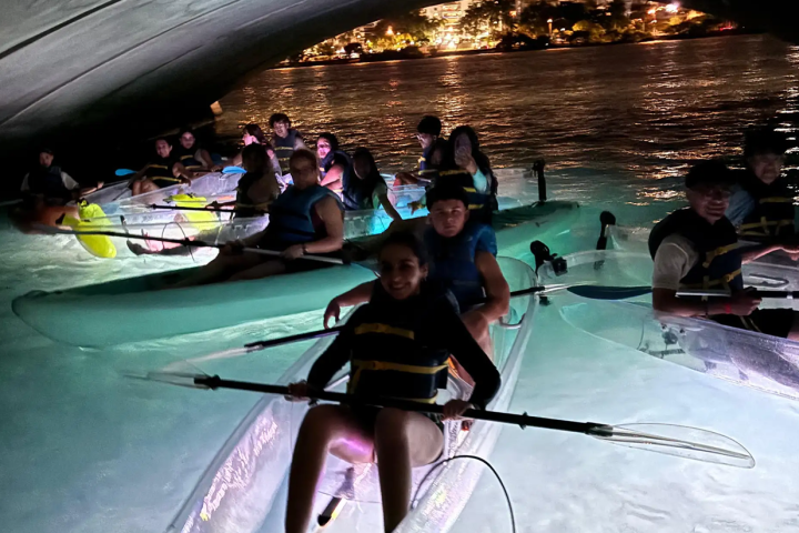 Group kayaking at night under a bridge with illuminated water.