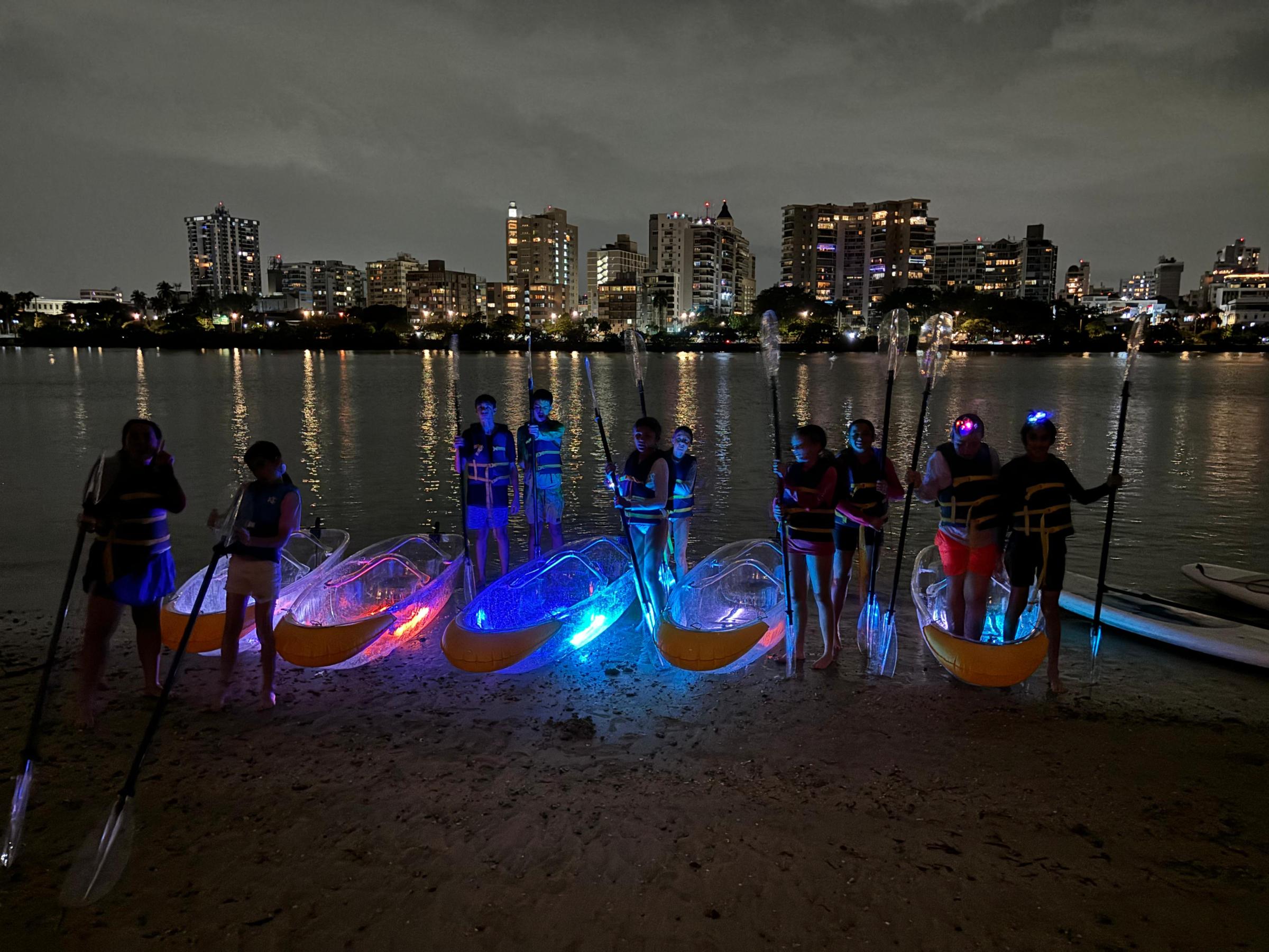 Group with illuminated kayaks by a city river at night.