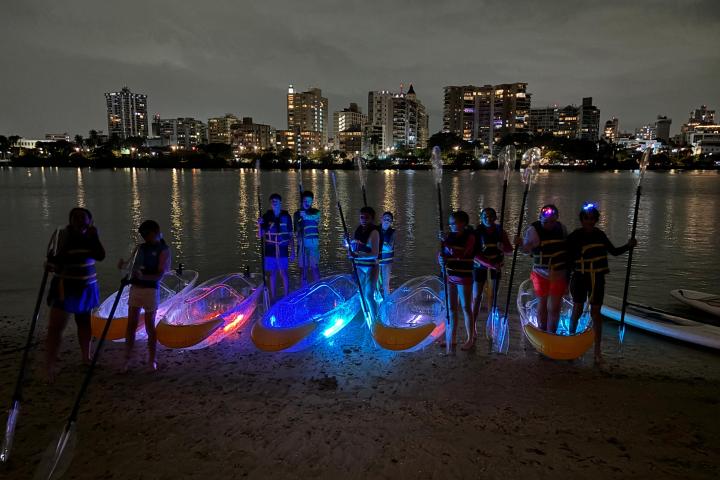 Group with illuminated kayaks by a city river at night.