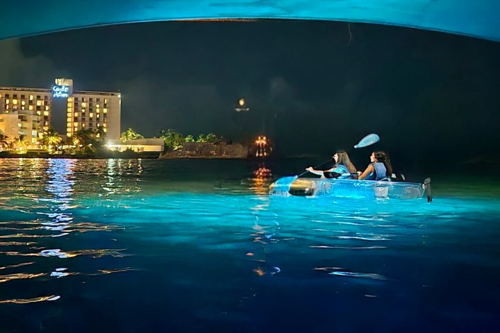 Two people kayaking at night near a lit-up building on the water.