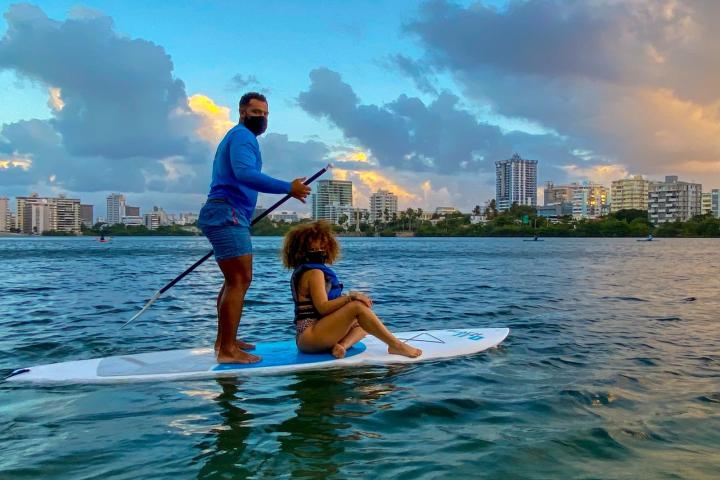 Two people on a paddleboard at sunset, with city skyline in the background.