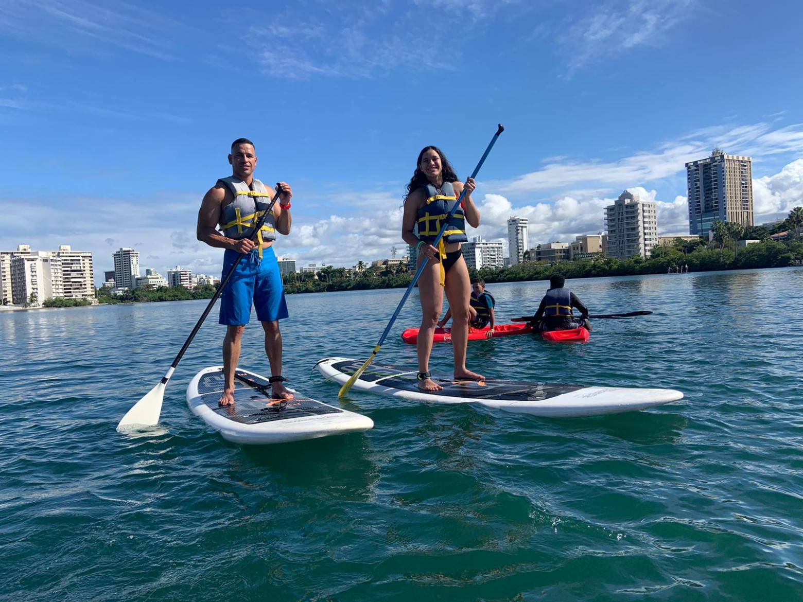 Two people paddleboarding on a calm waterway with city buildings in the background.