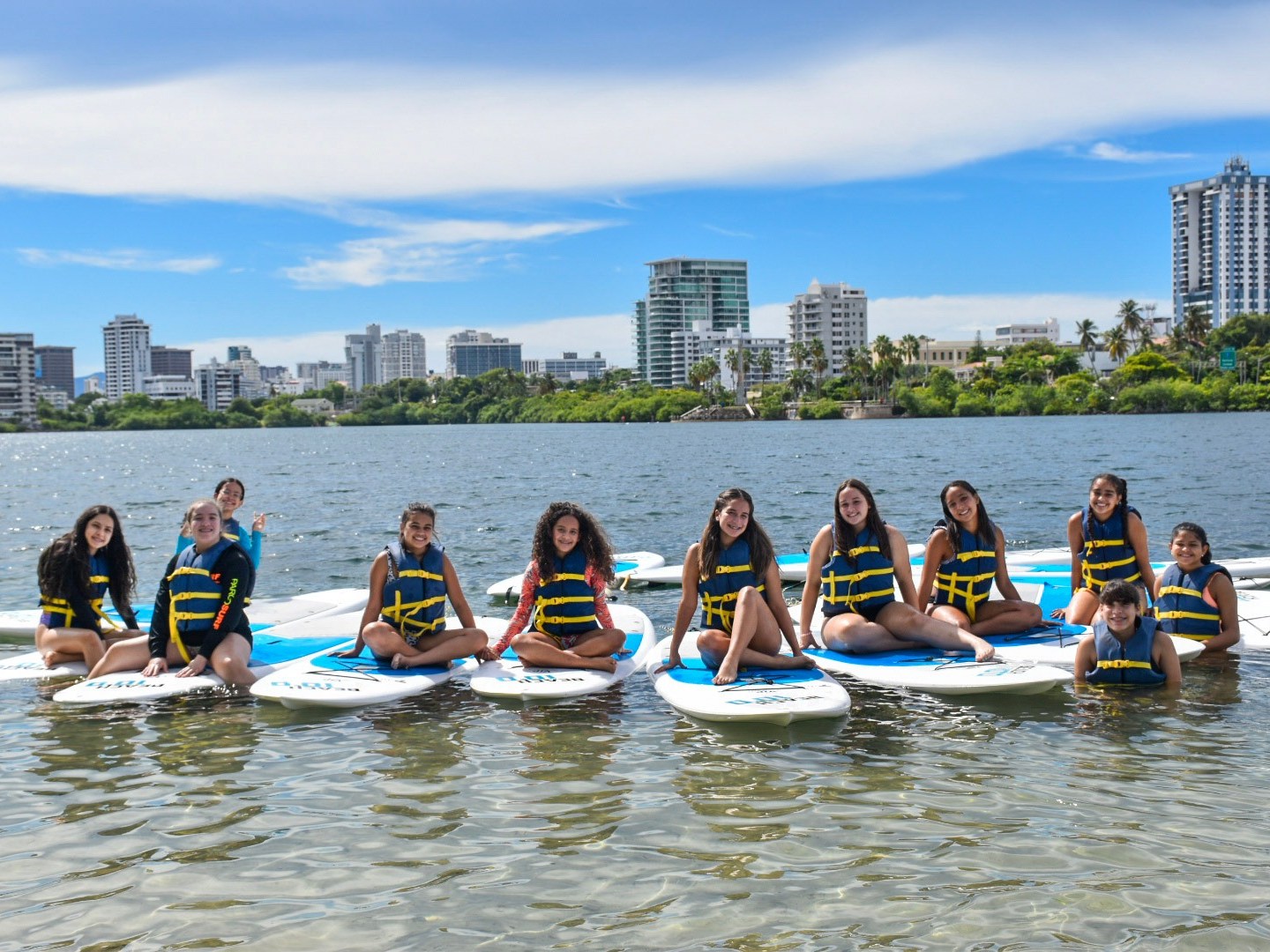 Group of young people wearing life vests sitting on paddleboards on a lake with city skyline in the background.