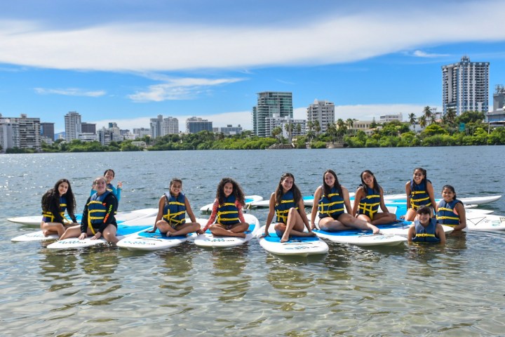 Group of young people wearing life vests sitting on paddleboards on a lake with city skyline in the background.