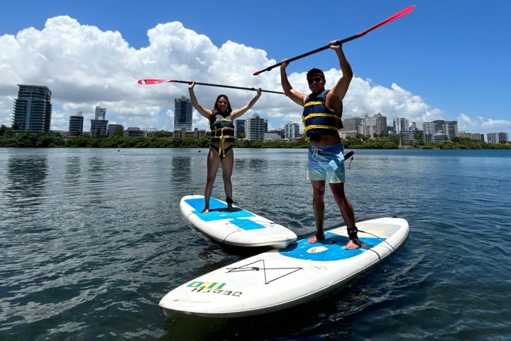 Two people on paddleboards raising paddles, with city skyline in background.