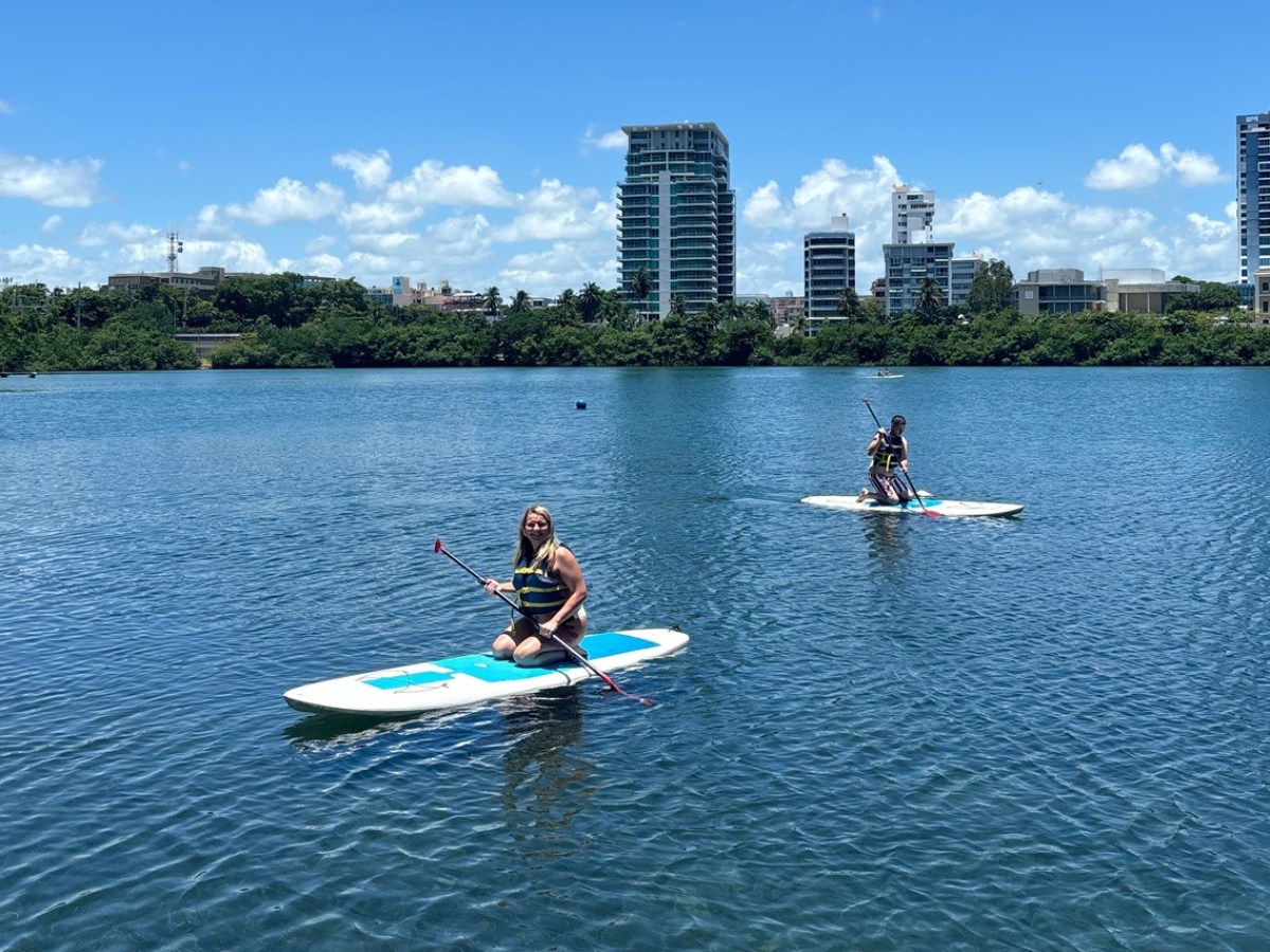 Two people paddleboarding on a calm lake with city buildings and trees in the background under a blue sky.
