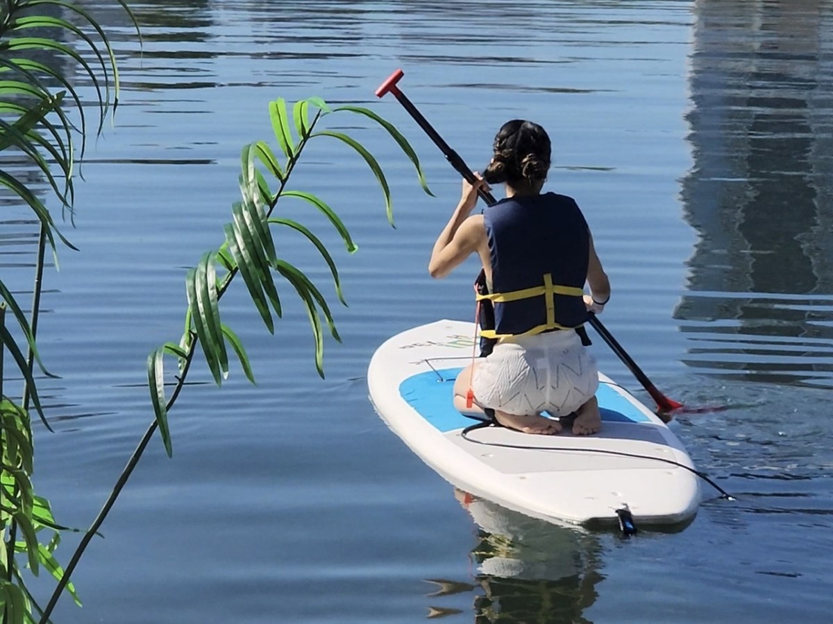 Person paddles on a stand-up paddleboard on a calm lake, wearing a life vest.