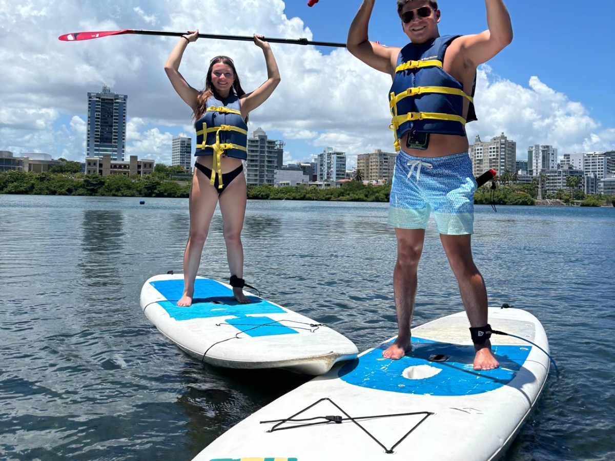 Two people in life jackets stand on paddleboards, holding paddles on a sunny day with city skyline in background.