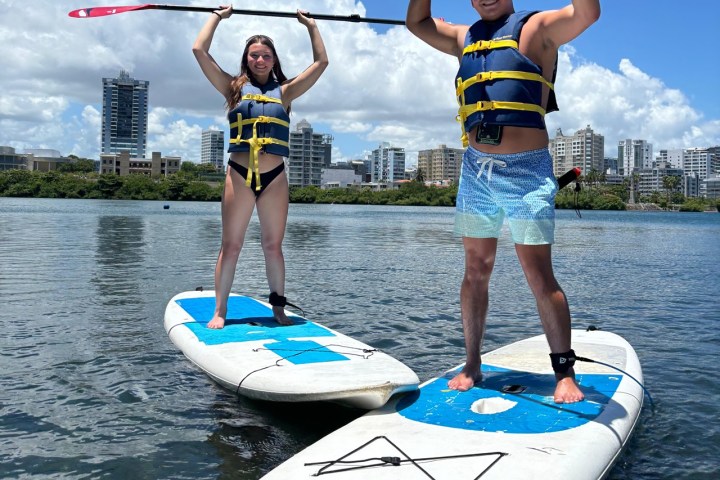 Two people in life jackets stand on paddleboards, holding paddles on a sunny day with city skyline in background.