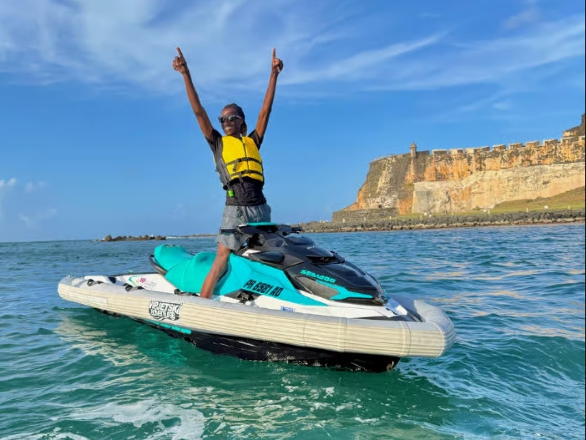 Person on jet ski raises arms in joy with an old fort and blue sky in the background.