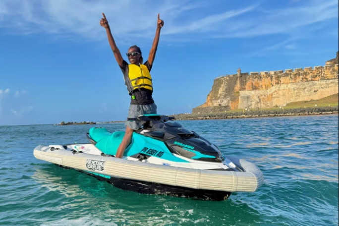 Person on jet ski raises arms in joy with an old fort and blue sky in the background.