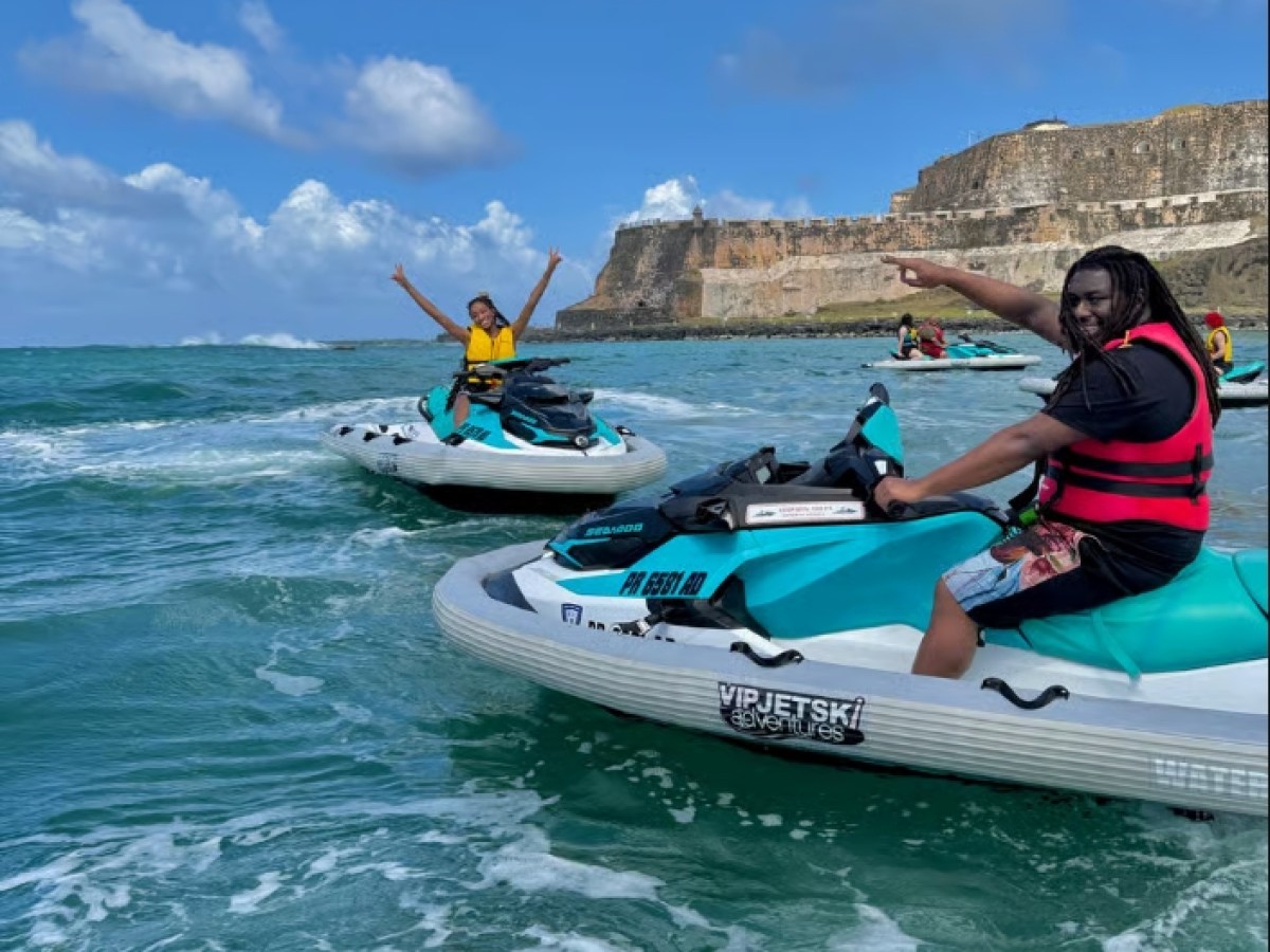 Two people on jet skis in ocean with a historical fort in the background, under a sunny sky.