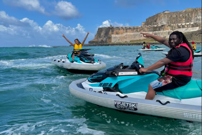 Two people on jet skis in ocean with a historical fort in the background, under a sunny sky.