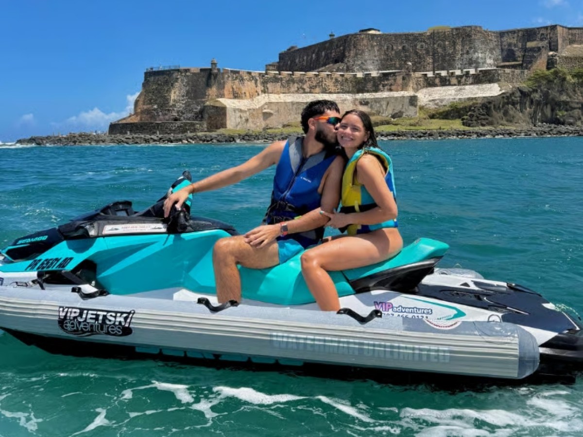 Couple on a jet ski kissing near a coastal fortress with blue sky and ocean.