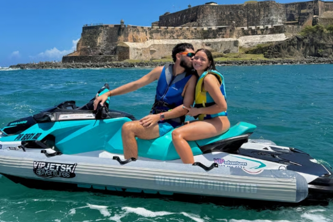 Couple on a jet ski kissing near a coastal fortress with blue sky and ocean.