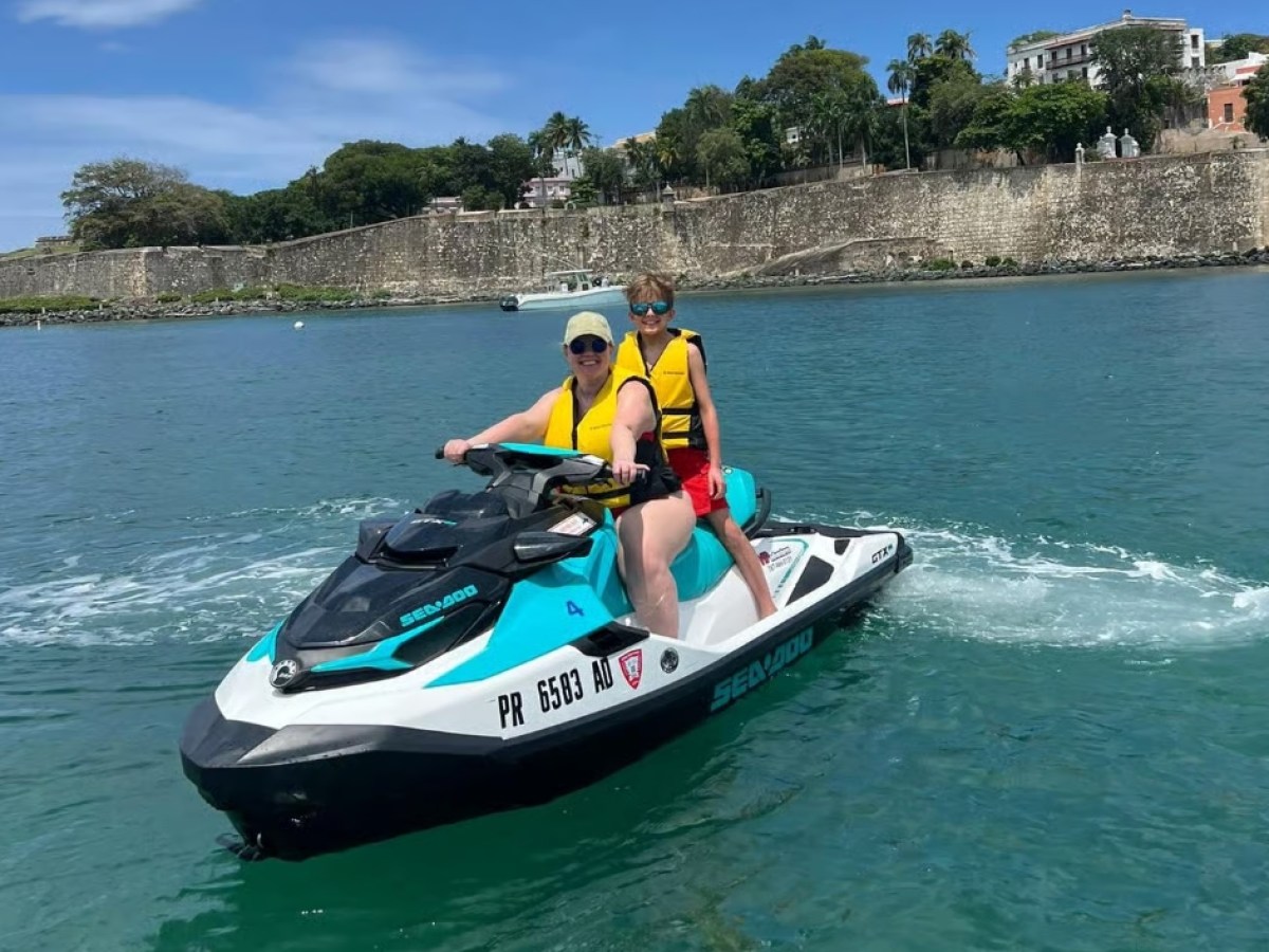 Two people on a jet ski in blue water with a stone wall and trees in the background.