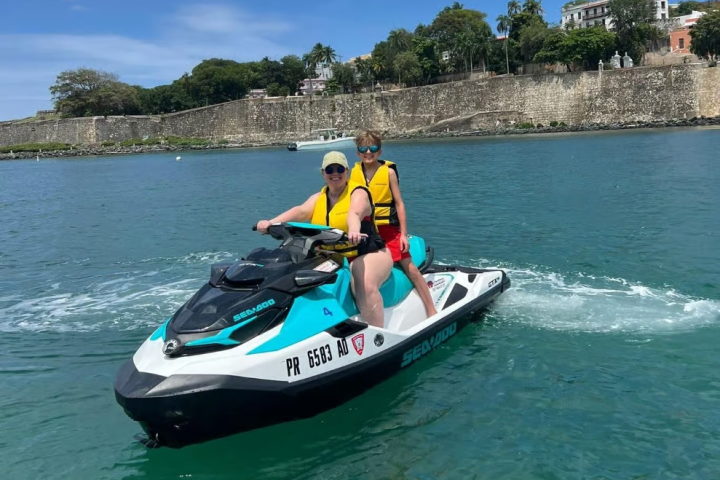 Two people on a jet ski in blue water with a stone wall and trees in the background.