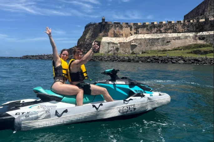Two people on a jet ski near a historic fort on a sunny day.