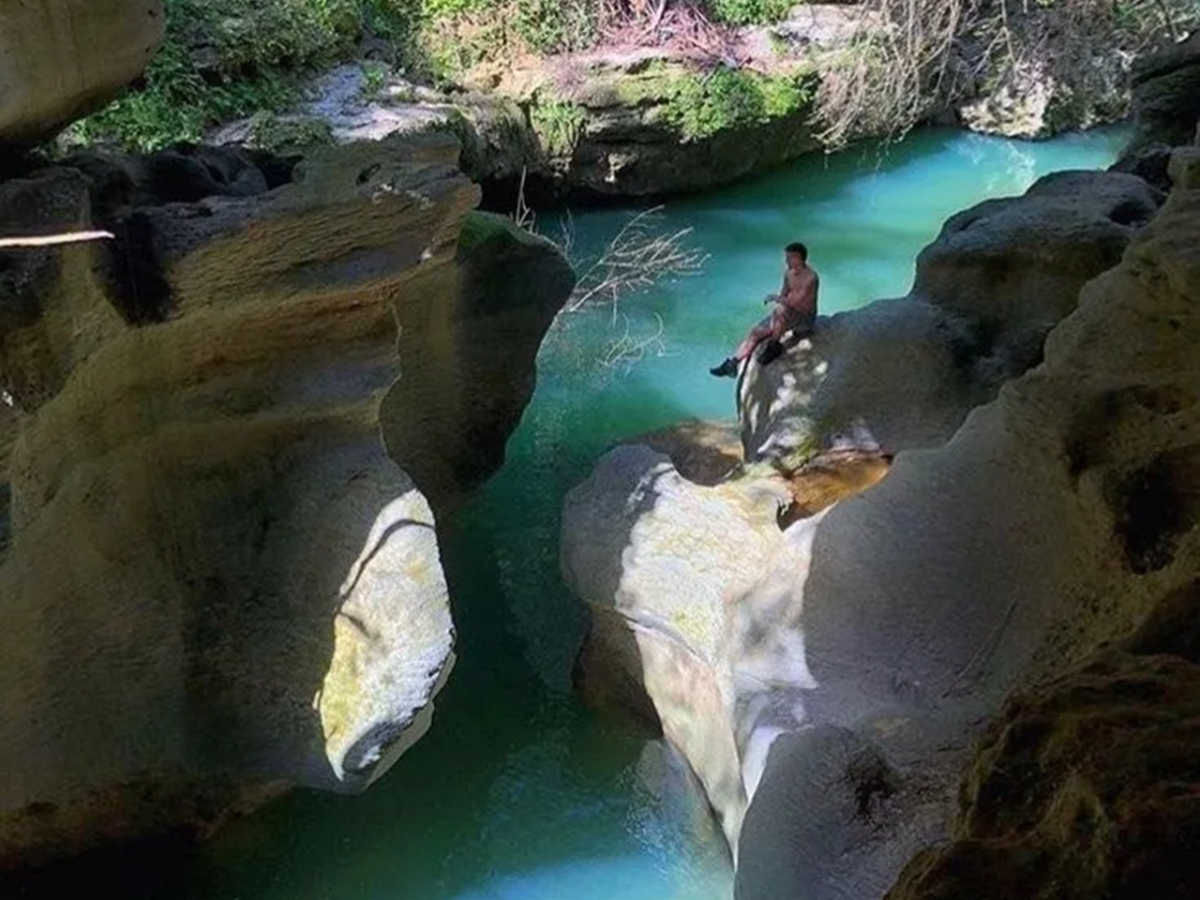 Person sitting on rock above turquoise water in a rocky cave opening.