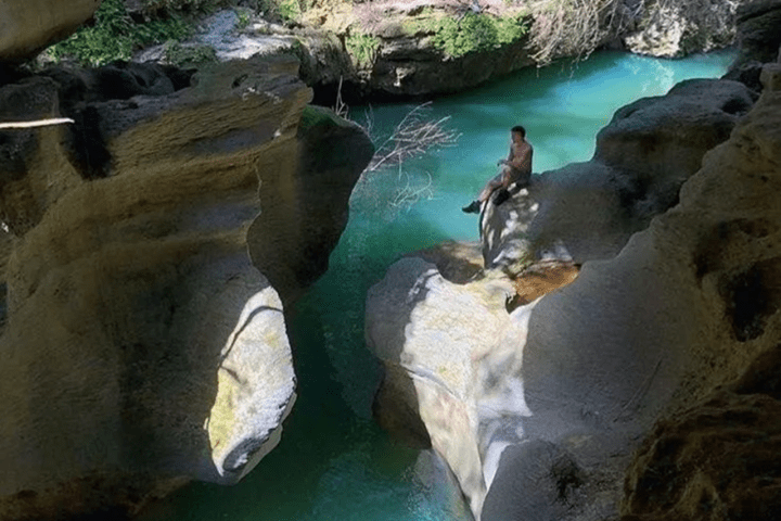 Person sitting on rock above turquoise water in a rocky cave opening.