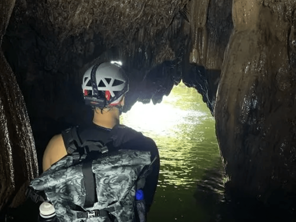 Person wearing helmet explores a dimly lit cave with water and rock formations.