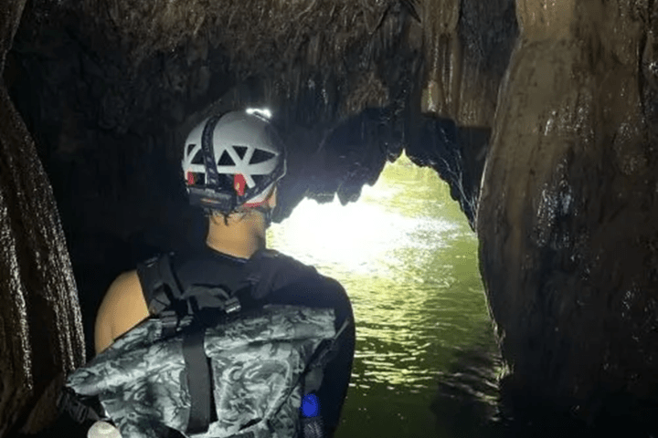 Person wearing helmet explores a dimly lit cave with water and rock formations.