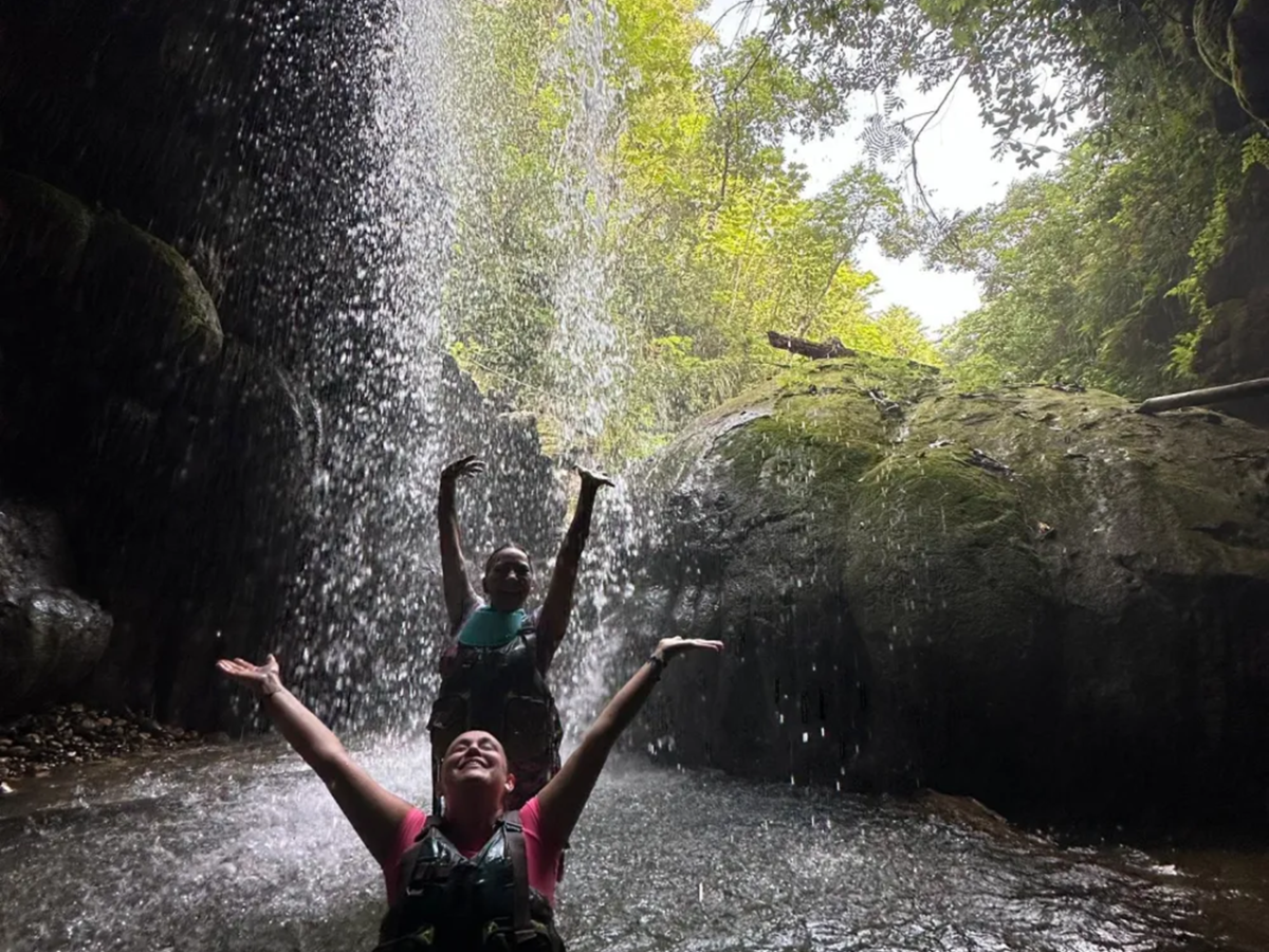Two people under a waterfall in a lush, rocky setting, enjoying the water.