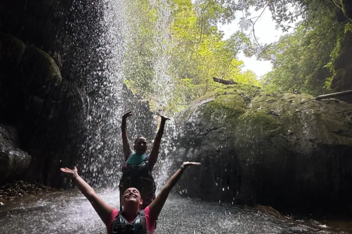 Two people under a waterfall in a lush, rocky setting, enjoying the water.