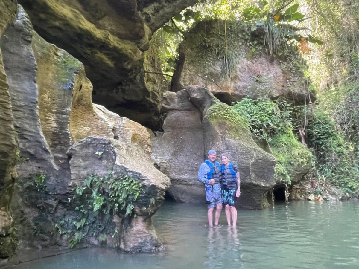 Two people stand in water by large rocks and greenery.