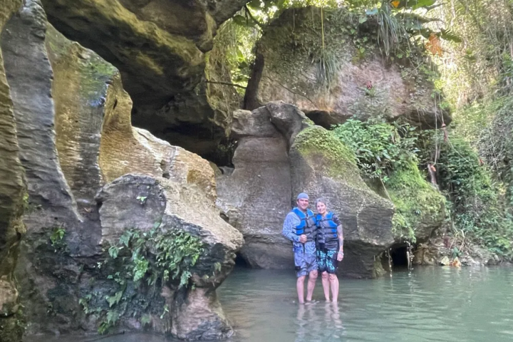 Two people stand in water by large rocks and greenery.