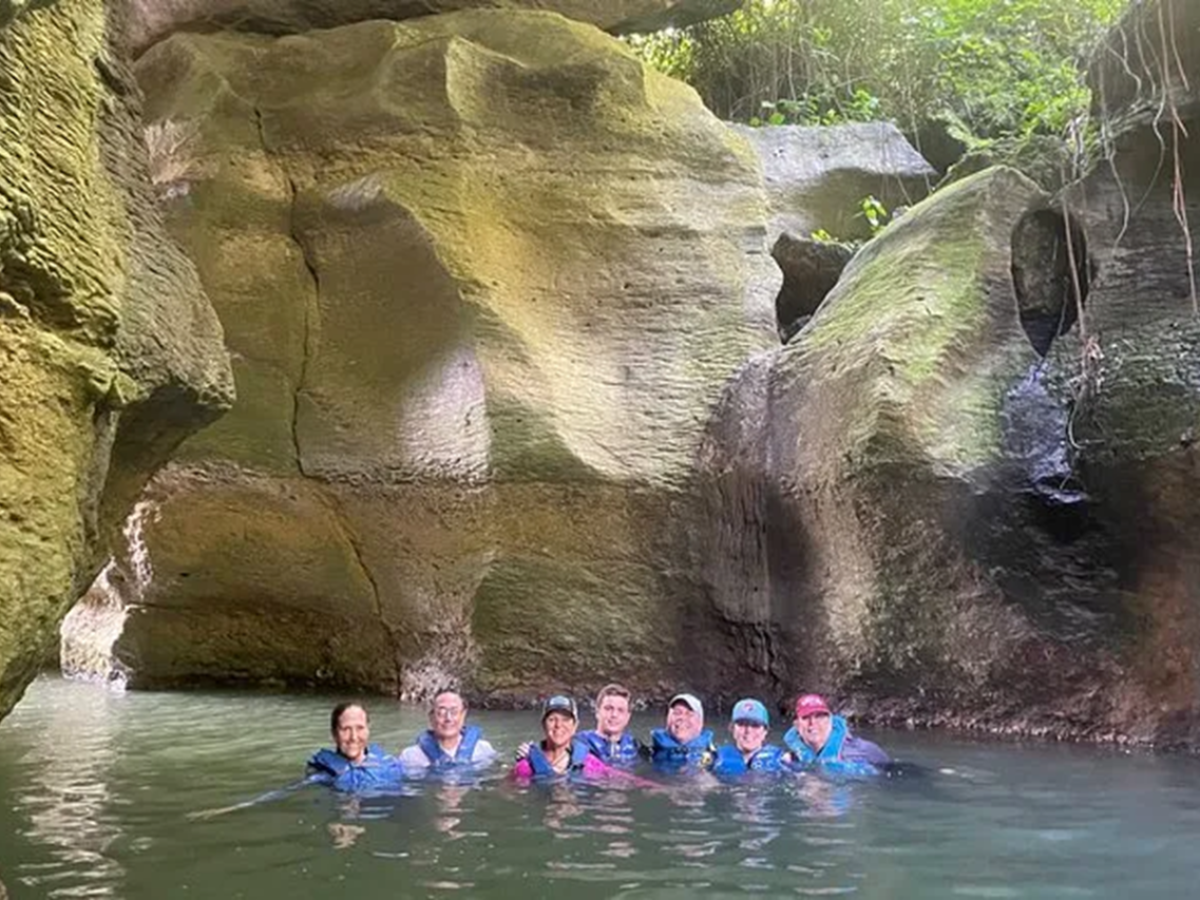 Group of seven people in a rocky cave pool, wearing life jackets.