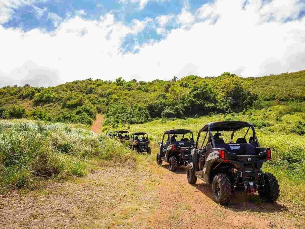 Four ATVs driving on a dirt path through grassy hills under a partly cloudy sky.