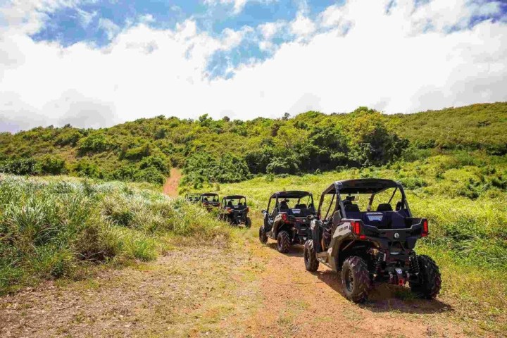 Four ATVs driving on a dirt path through grassy hills under a partly cloudy sky.