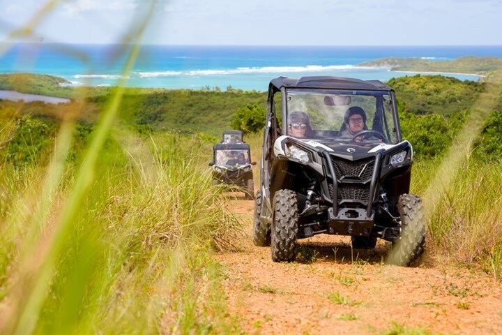 Two off-road vehicles driving on a dirt path with a scenic ocean view in the background.