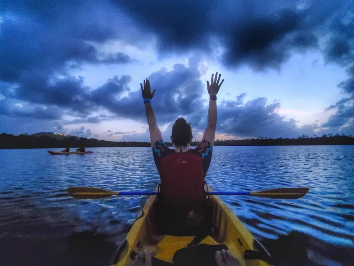 Person in kayak with arms raised, on a lake at dusk with clouded sky.
