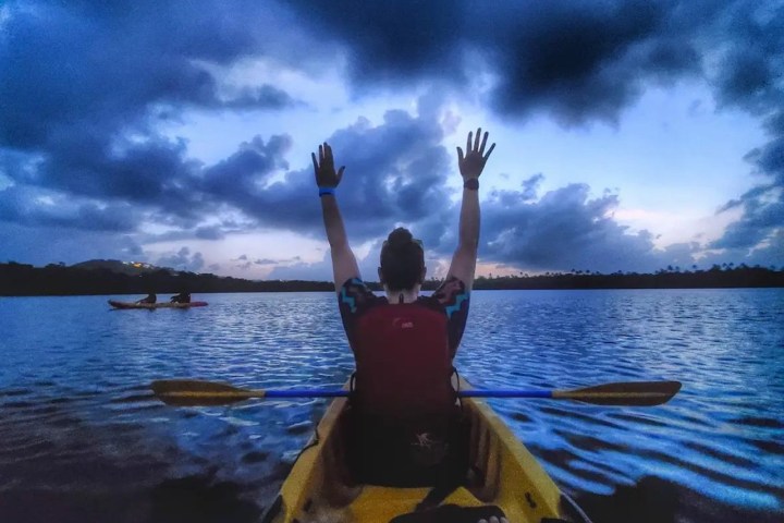 Person in kayak with arms raised, on a lake at dusk with clouded sky.
