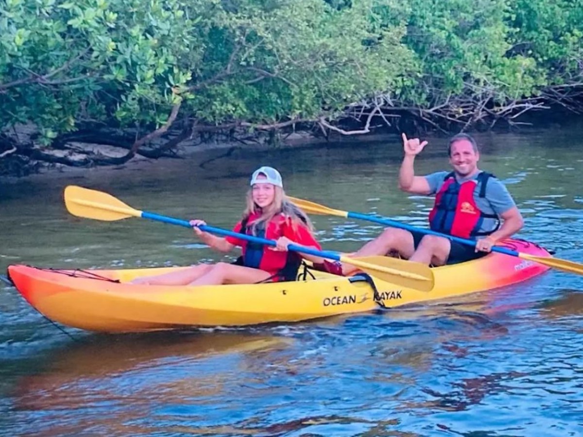 Two people in a kayak on a calm river, wearing life vests and holding paddles, near a leafy riverbank.
