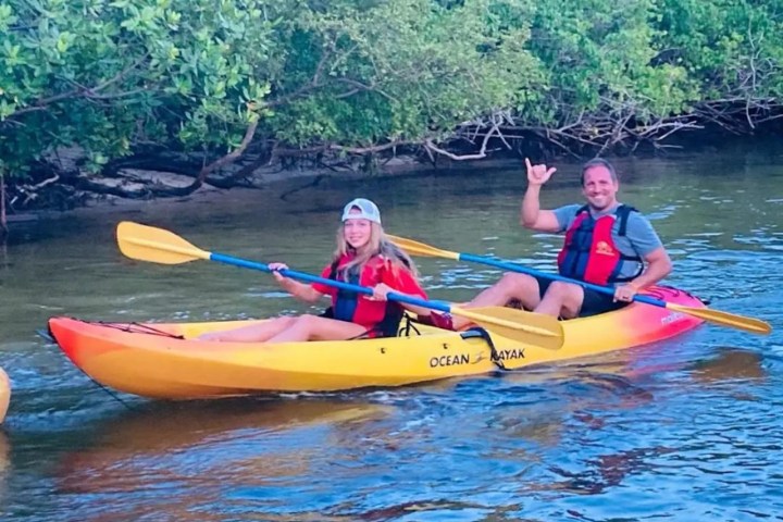 Two people in a kayak on a calm river, wearing life vests and holding paddles, near a leafy riverbank.