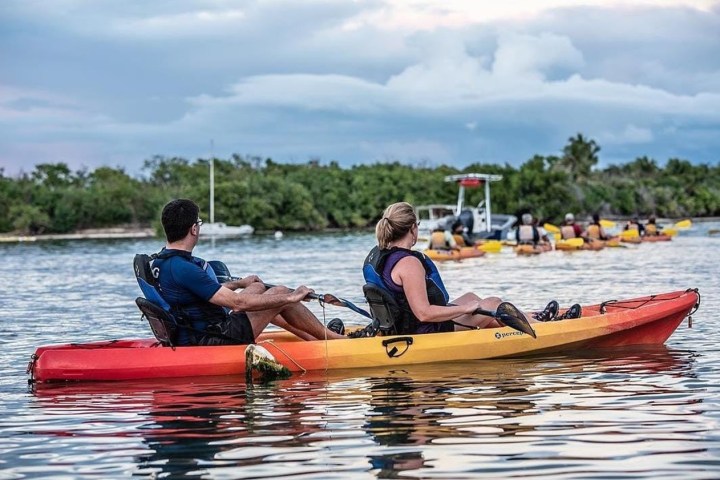 Two people in a tandem kayak on a calm river with others in the distance.