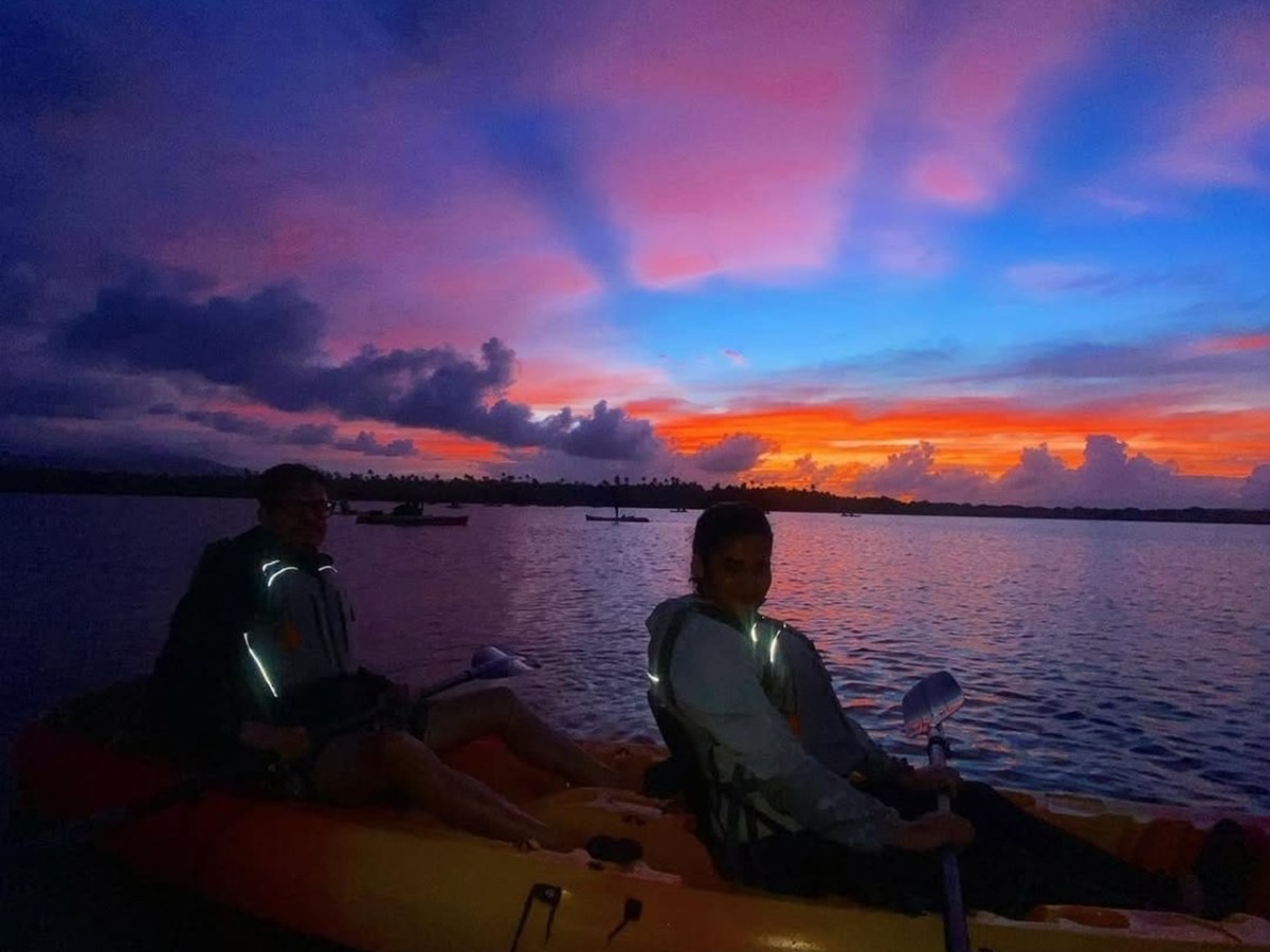 Two people kayaking at sunset with vibrant pink and blue sky reflected on water.
