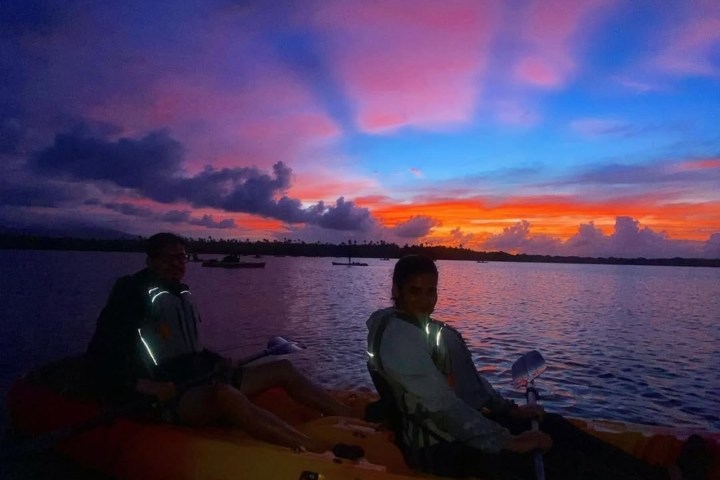 Two people kayaking at sunset with vibrant pink and blue sky reflected on water.