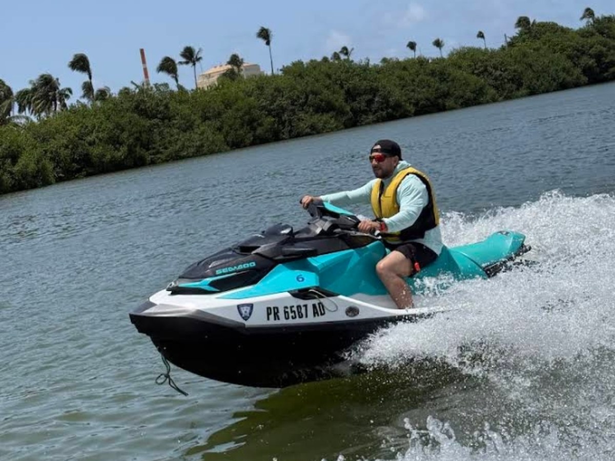 Person riding a jet ski wearing a yellow vest on a sunny day with trees in the background.