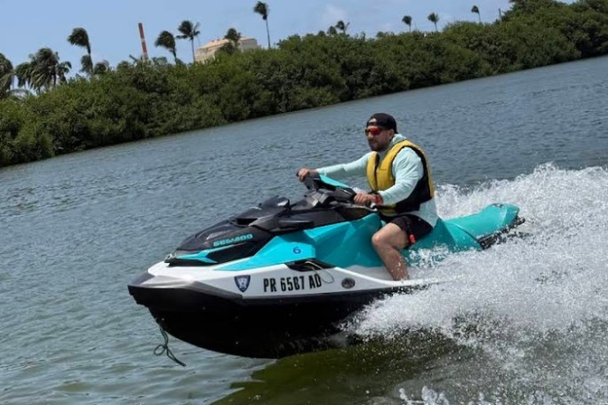 Person riding a jet ski wearing a yellow vest on a sunny day with trees in the background.