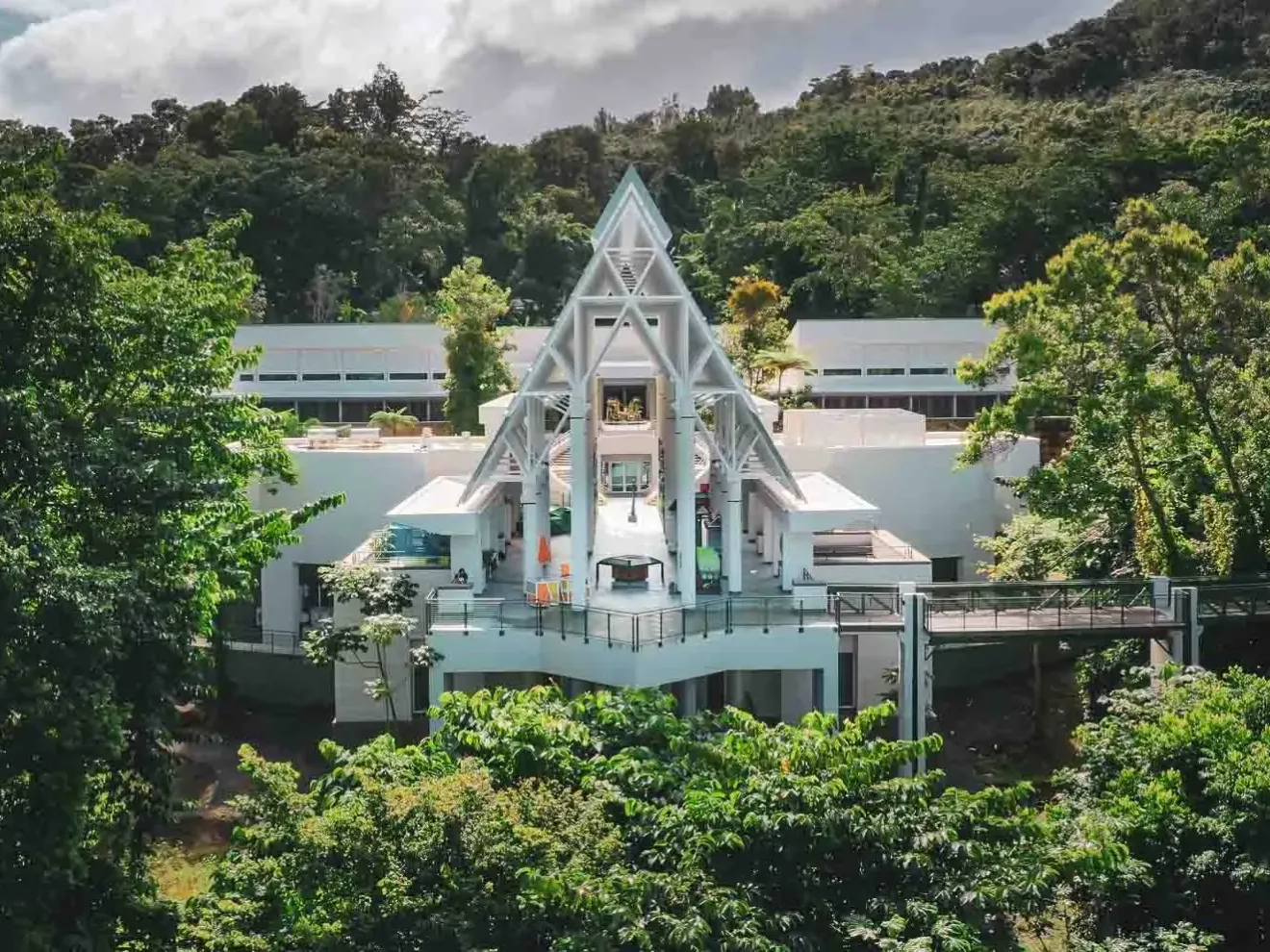 Modern white building with triangular roof surrounded by lush green trees.