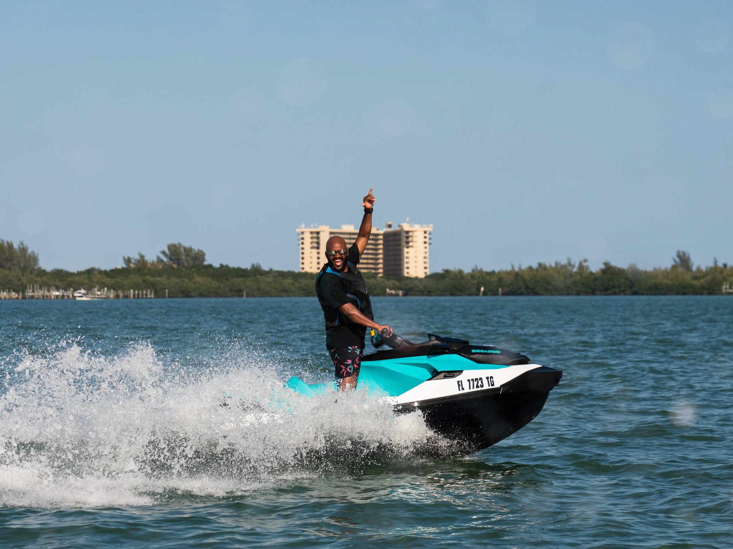 Person on a jet ski raising one arm, with buildings and trees in the background.