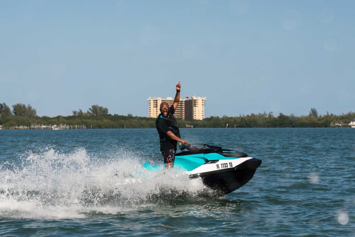 Person on a jet ski raising one arm, with buildings and trees in the background.