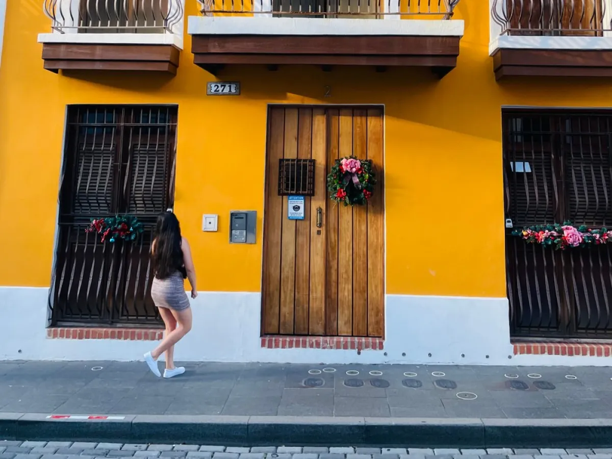 Person walking by a yellow building with wooden doors and floral wreath decorations.
