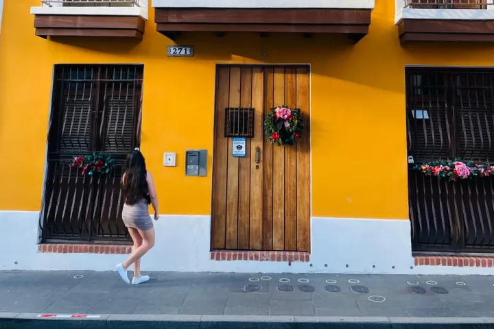 Person walking by a yellow building with wooden doors and floral wreath decorations.