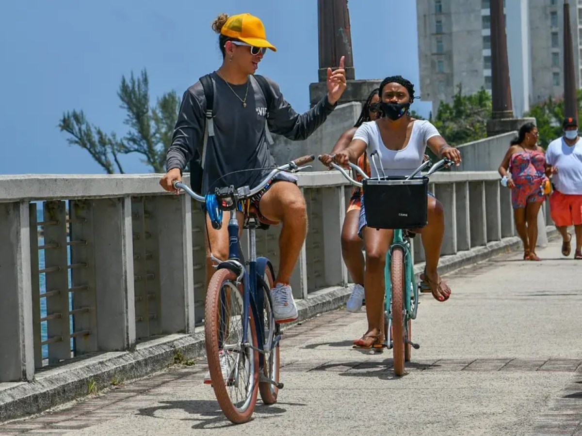Two people biking on a boardwalk, one wearing a yellow cap, others walking in the background.