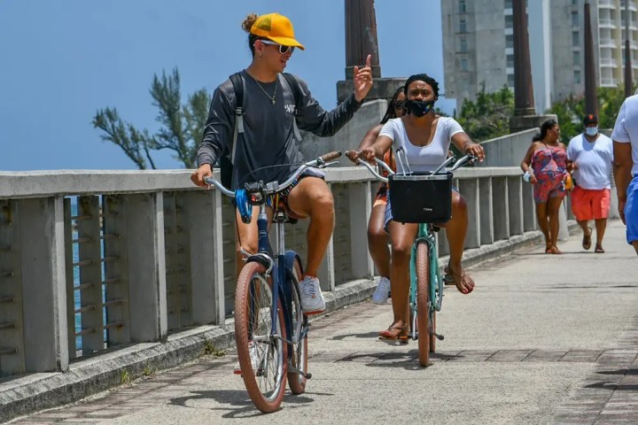 Two people biking on a boardwalk, one wearing a yellow cap, others walking in the background.